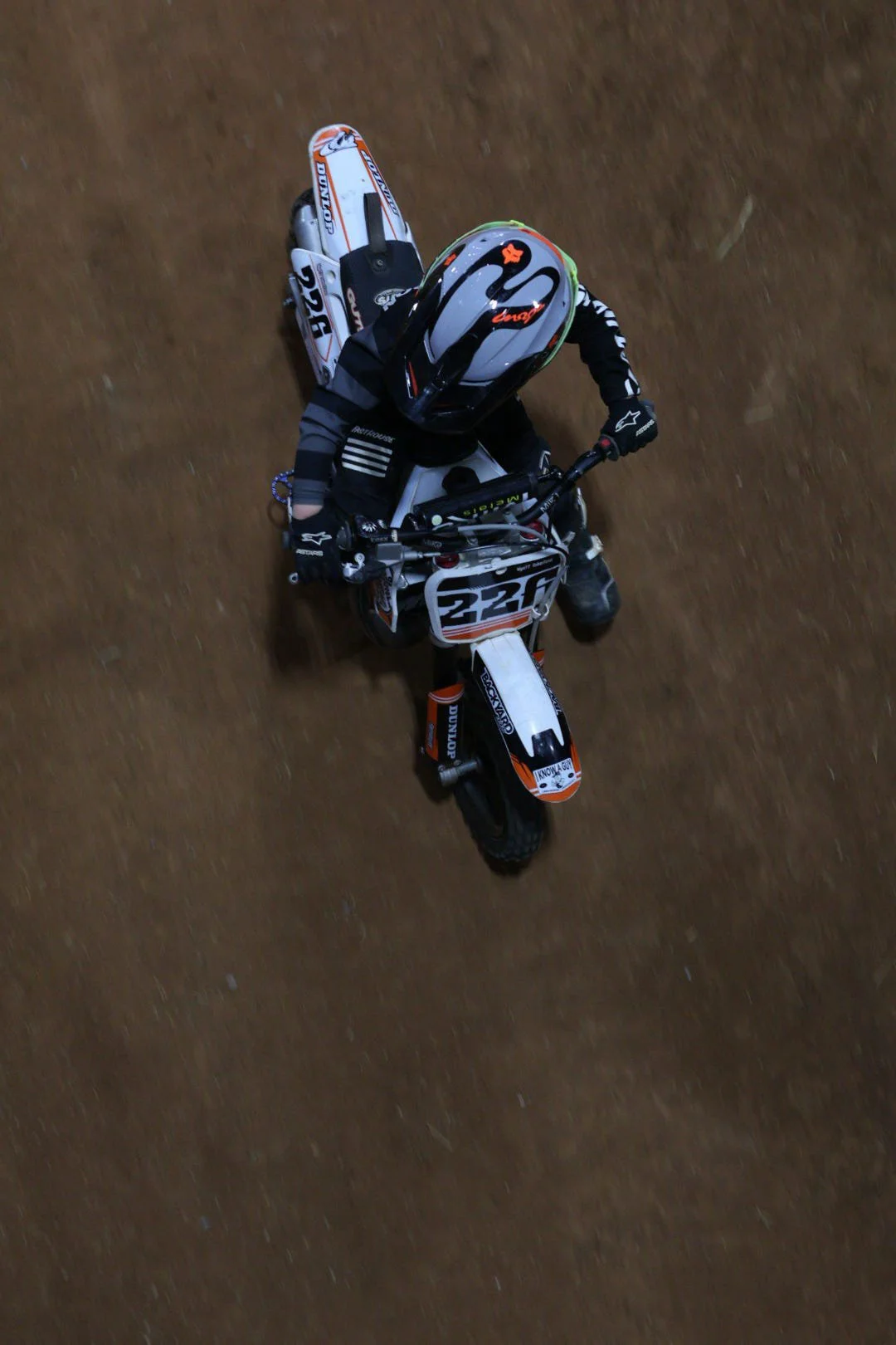 Top-down view of a motocross rider on a dirt track, wearing a helmet and black riding gear, riding a dirt bike numbered 220 with orange, white, and black decals.