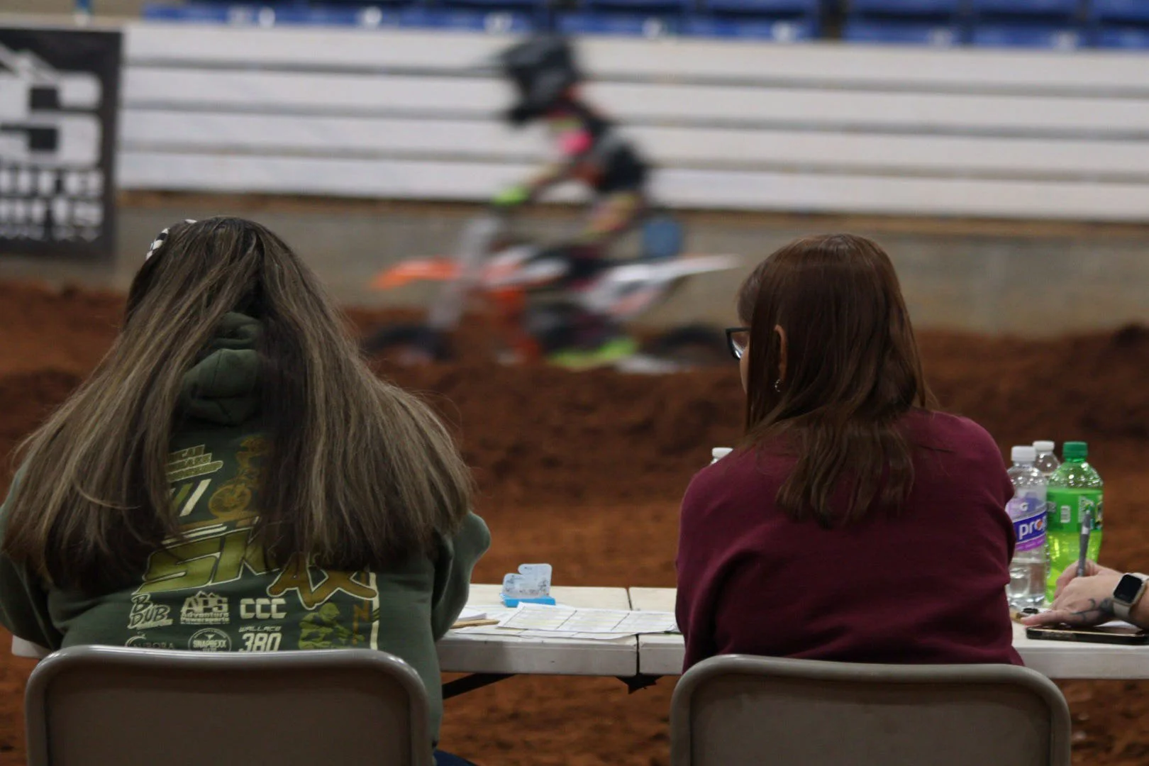 Two women seated at a table watching a dirt bike race in an indoor arena, with water bottles on the table.