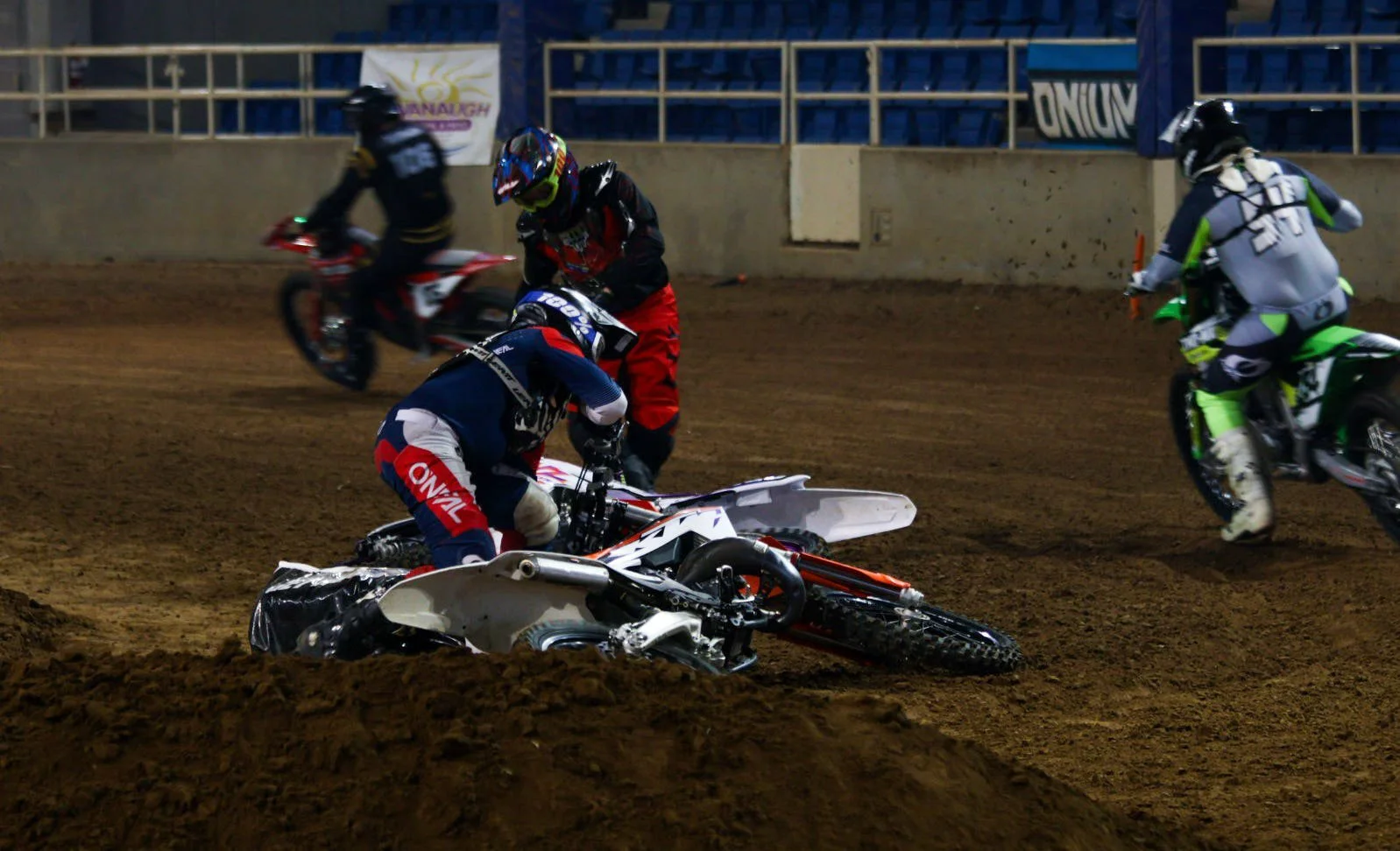 A motocross accident scene with a fallen rider and other riders around on a dirt track at night.