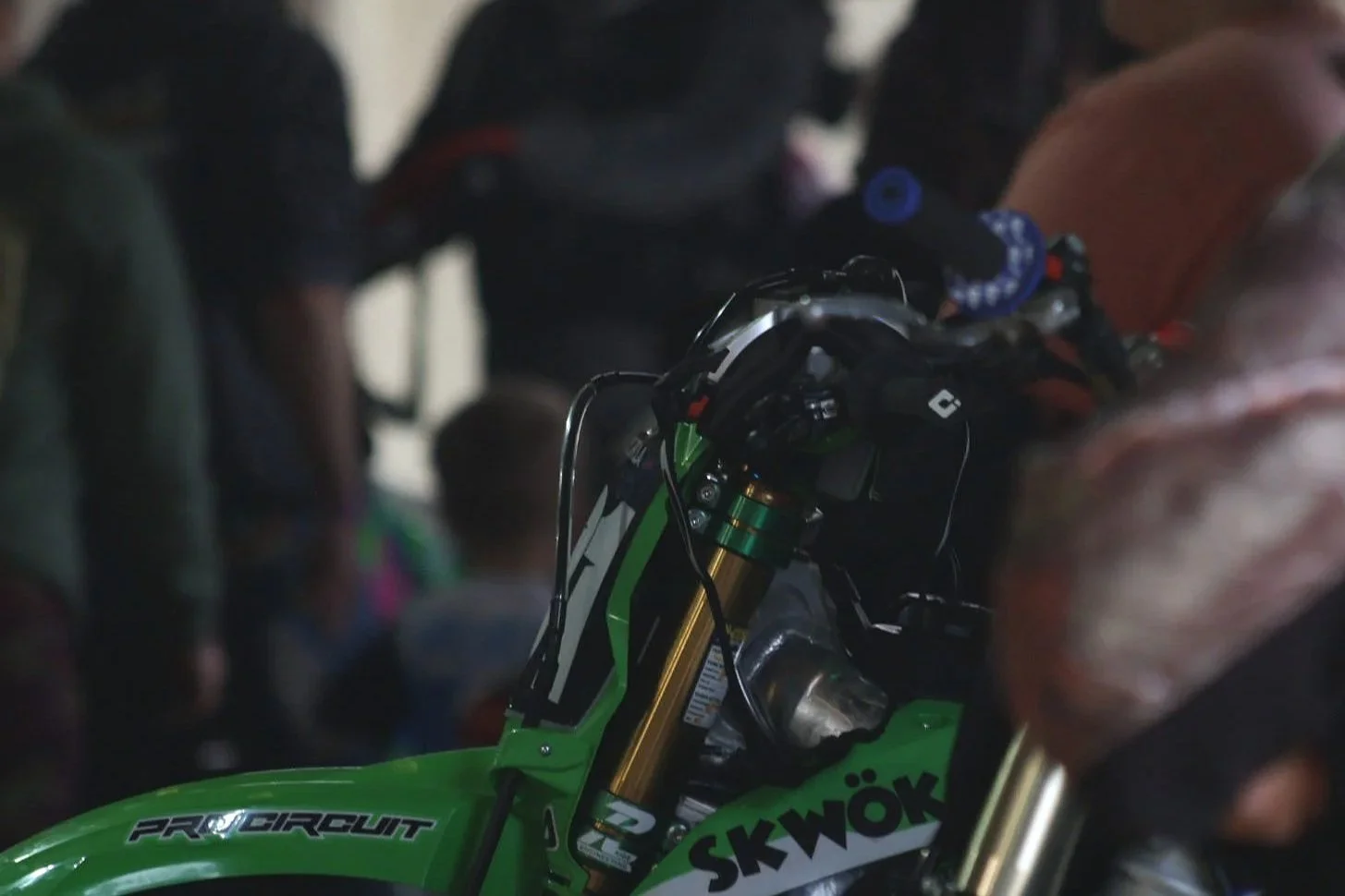 Close-up of a green motocross bike with gold suspension forks, surrounded by people at an event or gathering.