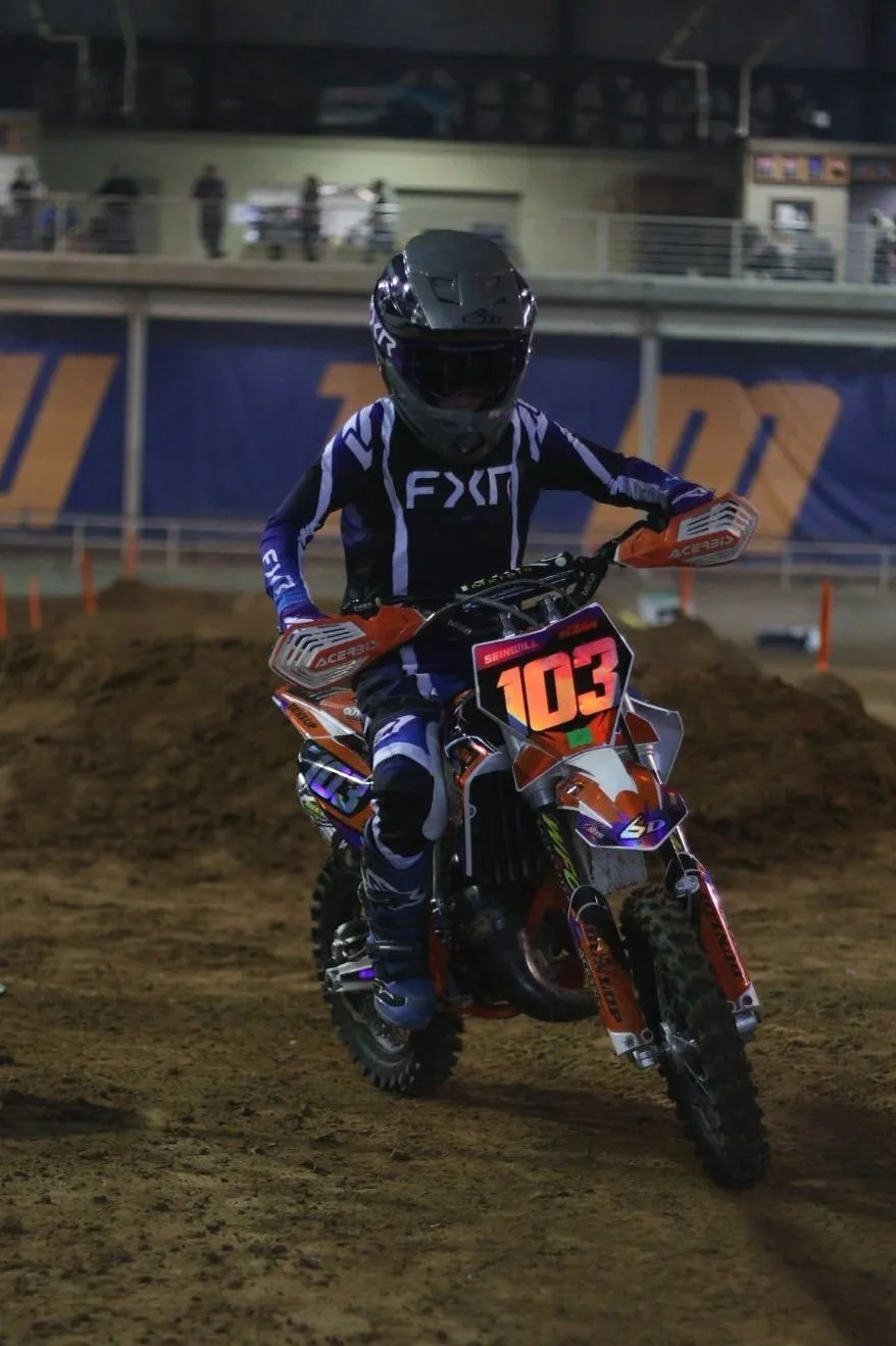 Motorcyclist riding on a dirt track at night, wearing full gear and helmet, with the number 103 on the front of the bike.