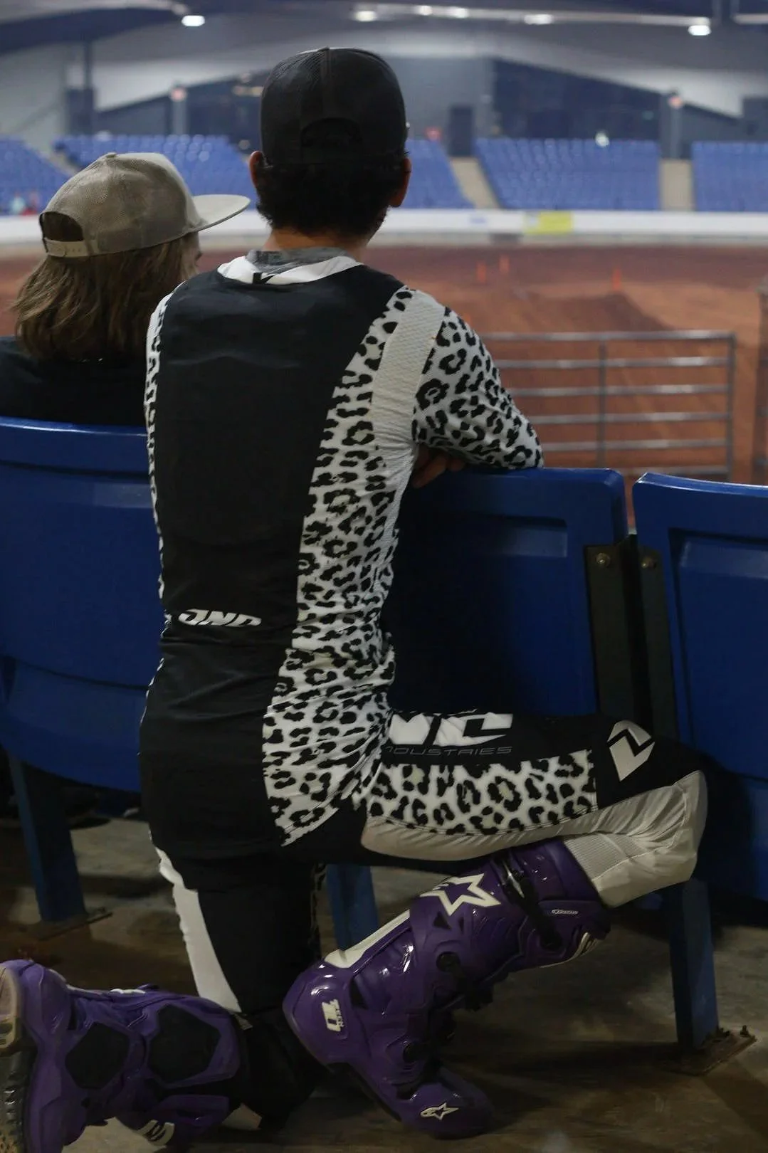 Two people, one wearing a black and white animal print outfit and a black cap, sitting in a stadium watching an indoor dirt bike or motocross event. The person on the right has purple motocross boots and increase ankle protection. The stadium has blu