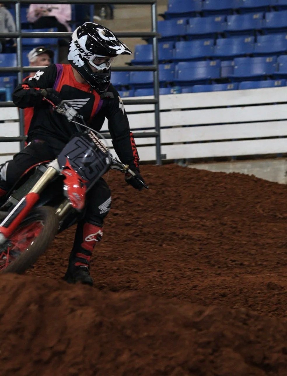 An individual riding a dirt bike on a dirt track in an indoor arena, wearing a helmet, riding gear, and gloves.
