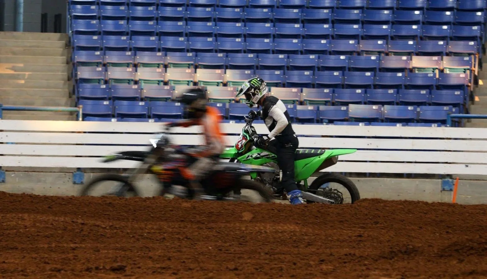 A person riding a green dirt bike on a dirt track, with another rider blurred in motion in front, in an indoor stadium with blue empty seats in the background.