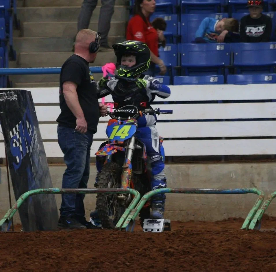 A young dirt bike rider wearing a helmet and racing gear on a dirt track, talking to an adult man in black T-shirt and jeans, inside an indoor arena with spectators in the background.