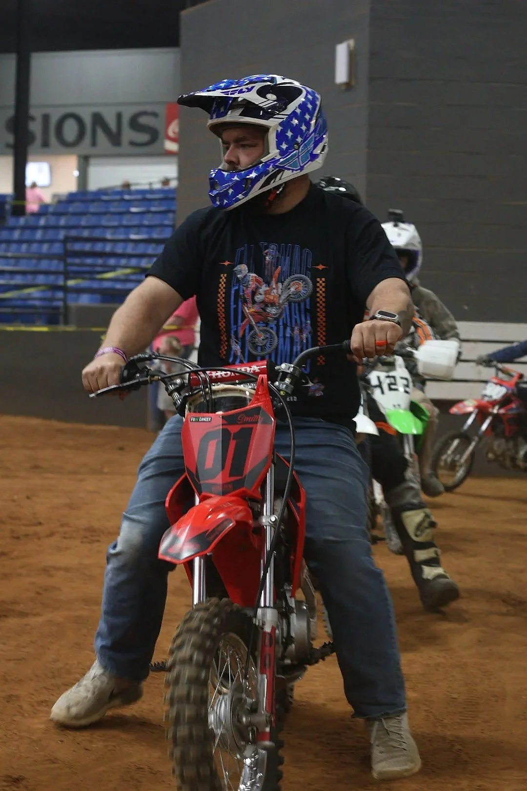 A young man riding a red and black dirt bike inside a racing arena, wearing a blue and white helmet and a black T-shirt with a motorcycle graphic, with other riders and bikes in the background.