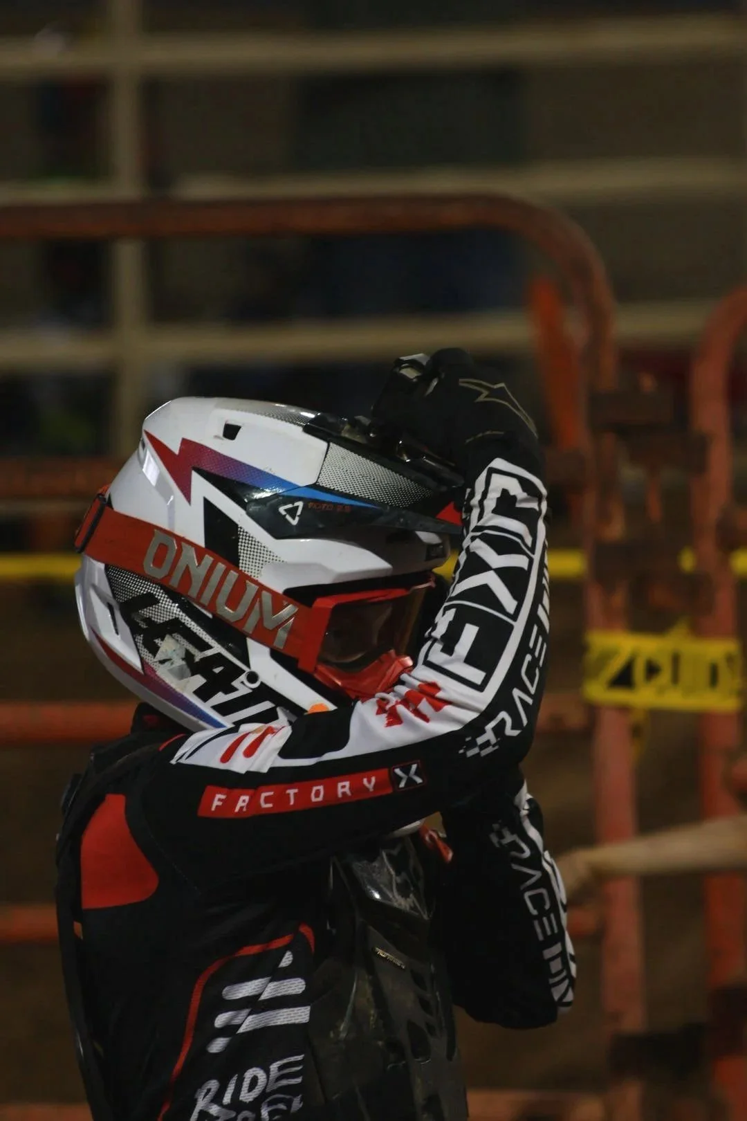 A motocross rider in black, white, red, and orange gear holding a motocross helmet, standing near a dirt track with orange fencing and yellow caution tape in the background.
