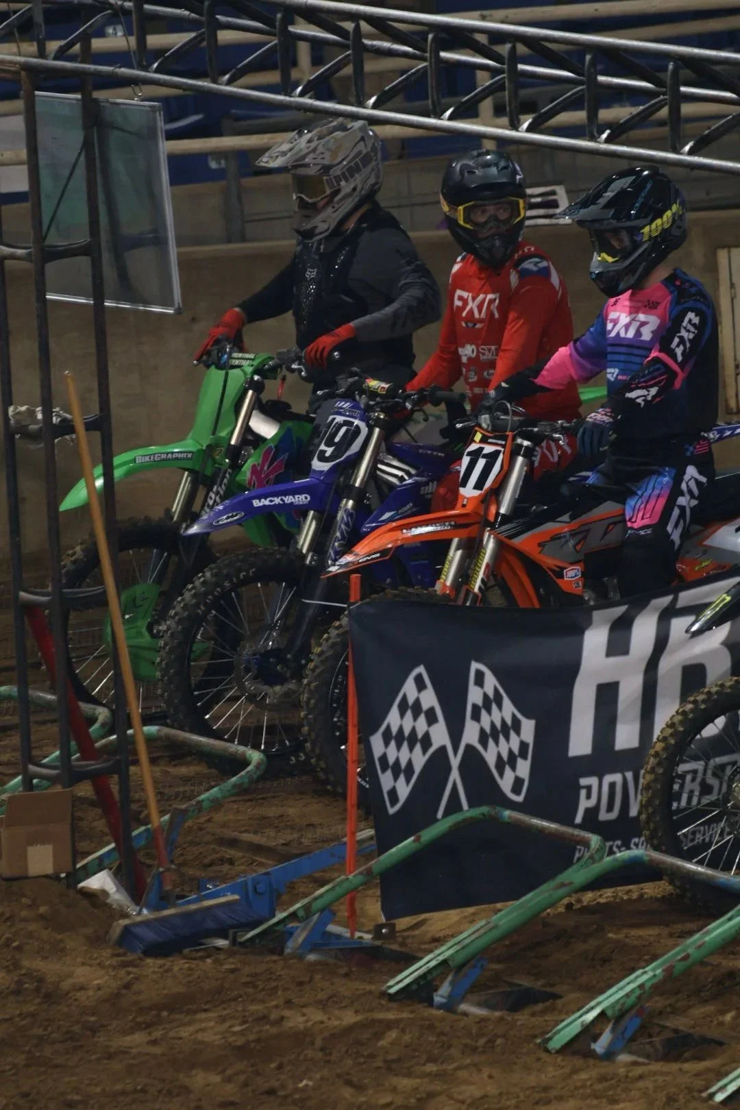 Three motocross riders wearing helmets and gear, sitting on their dirt bikes at a motocross track, with a banner and scaffolding nearby.