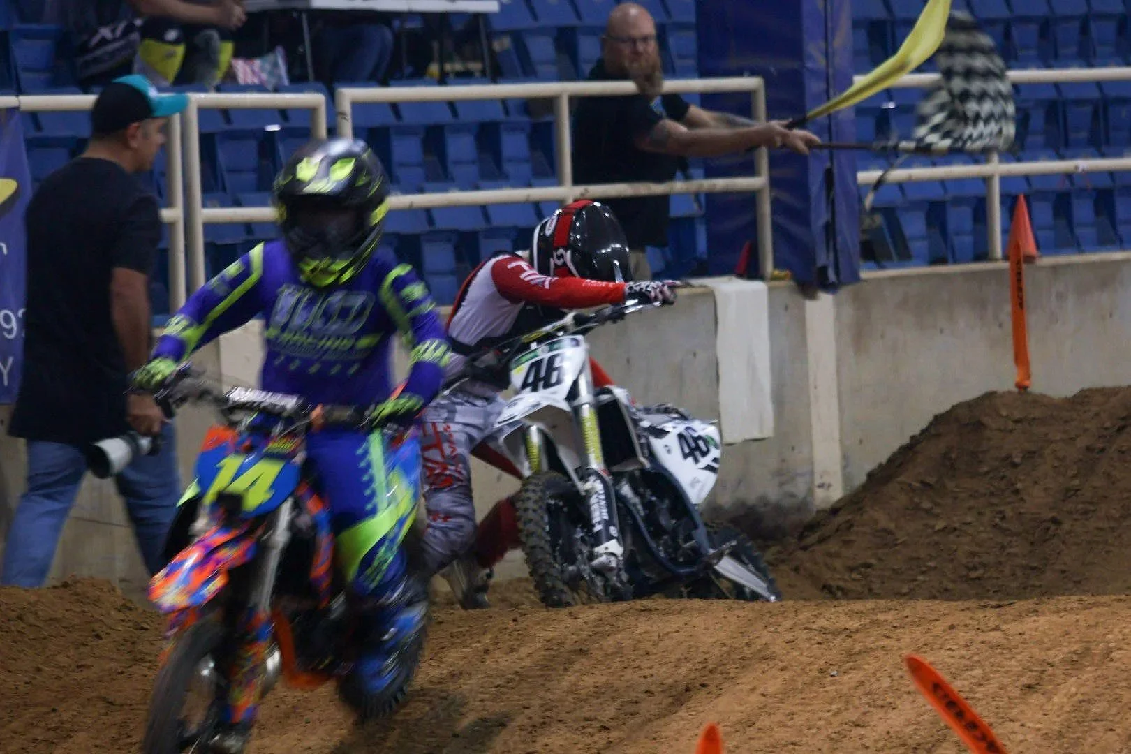 Two motocross riders and a mechanic on a dirt track in an indoor stadium. One rider is in blue and green gear, and the other is in red and white gear, entering a turn. A man with a beard and glasses is standing behind the concrete barrier, waving a y