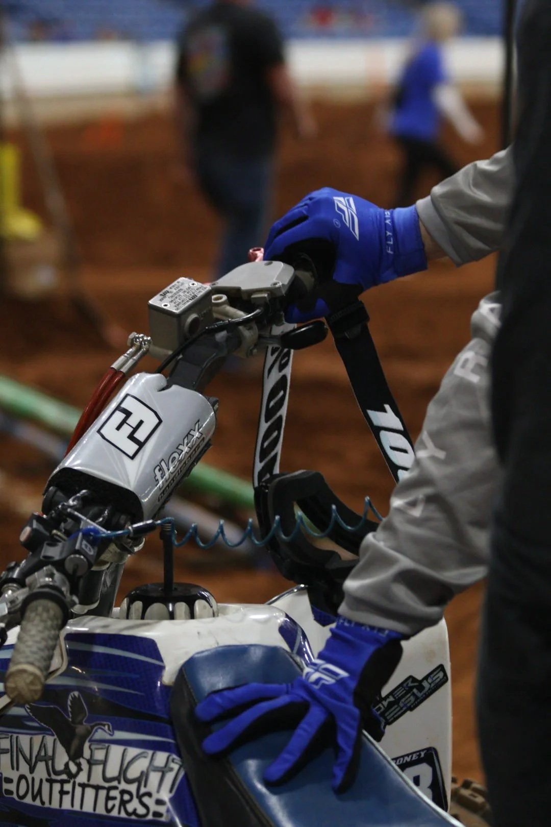 A person wearing blue gloves and holding the handlebar of a dirt bike, with a blurred indoor dirt track and other people in the background.