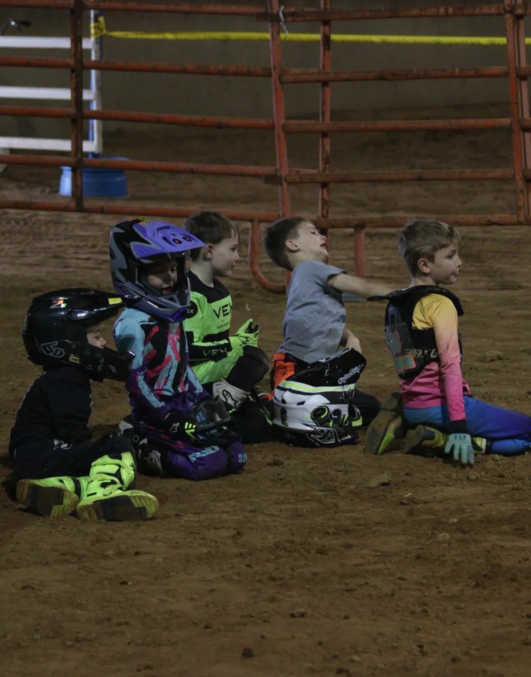 Five children kneeling on a dirt indoor dirt track, wearing motocross helmets and gear, with some pulling up and some kneeling.