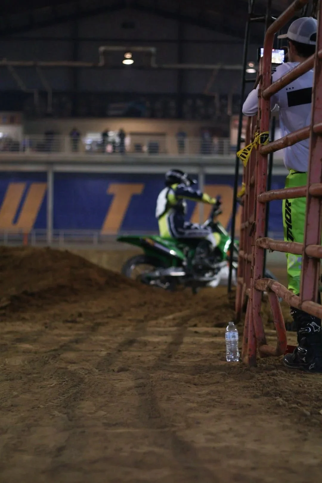 A person standing behind a metal barrier with a dirt bike in the background, inside a stadium or indoor arena. The scene appears to be at a motocross or dirt bike event.