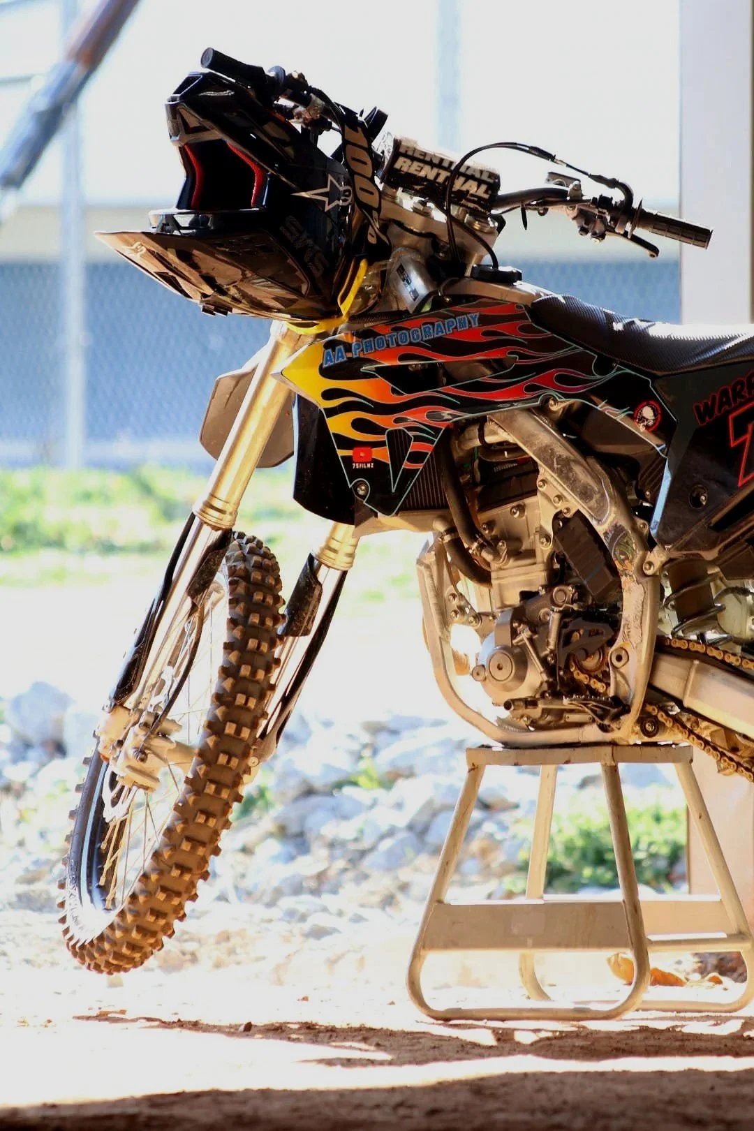 A dirt bike with a black helmet hanging from the handlebars, featuring colorful flame decals and various stickers, resting on a small stand on a dirt ground with rocks and grass in the background.