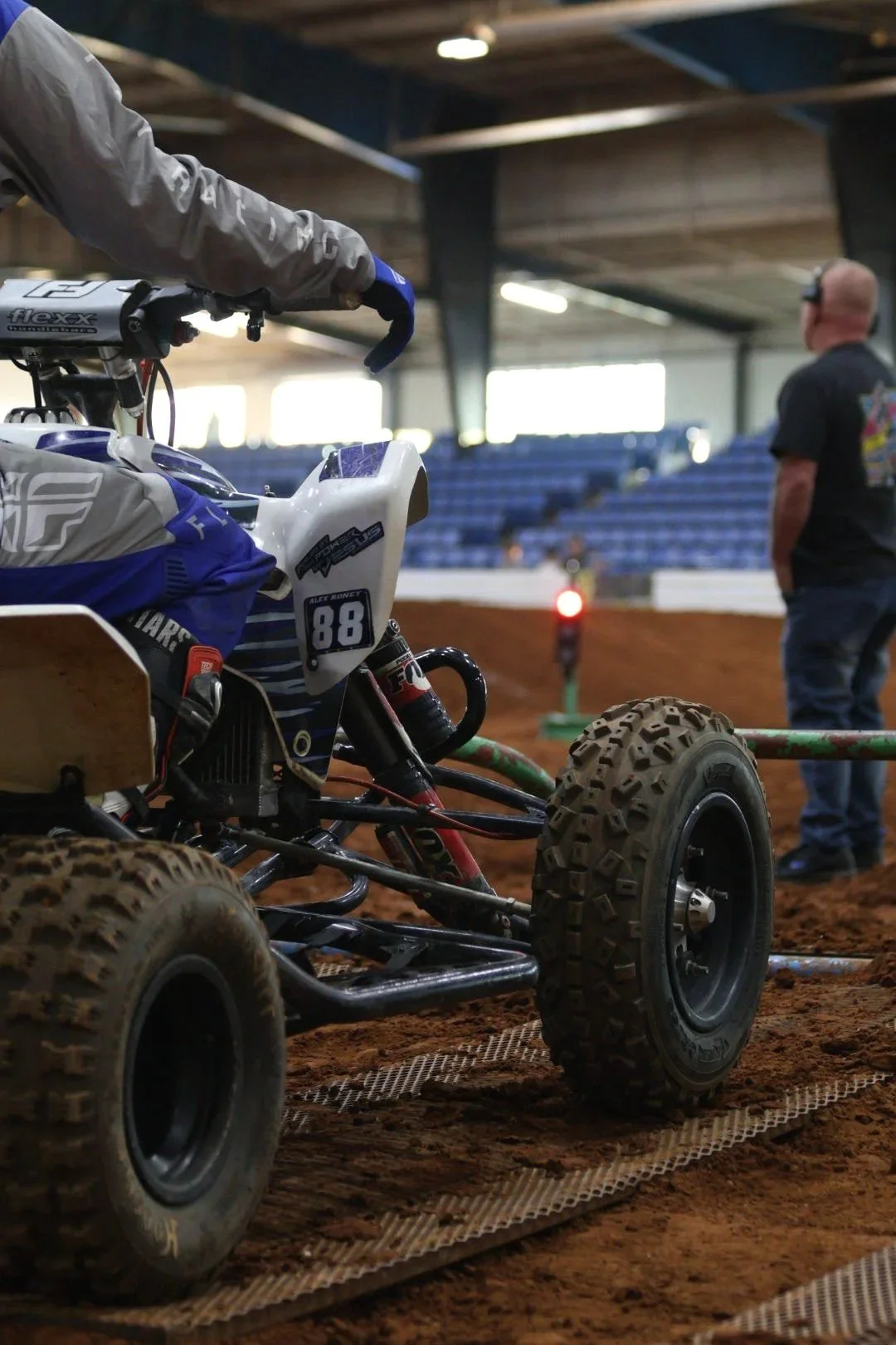 Close-up of an off-road racing vehicle with large tires on a dirt track inside a building, with a person standing in the background.