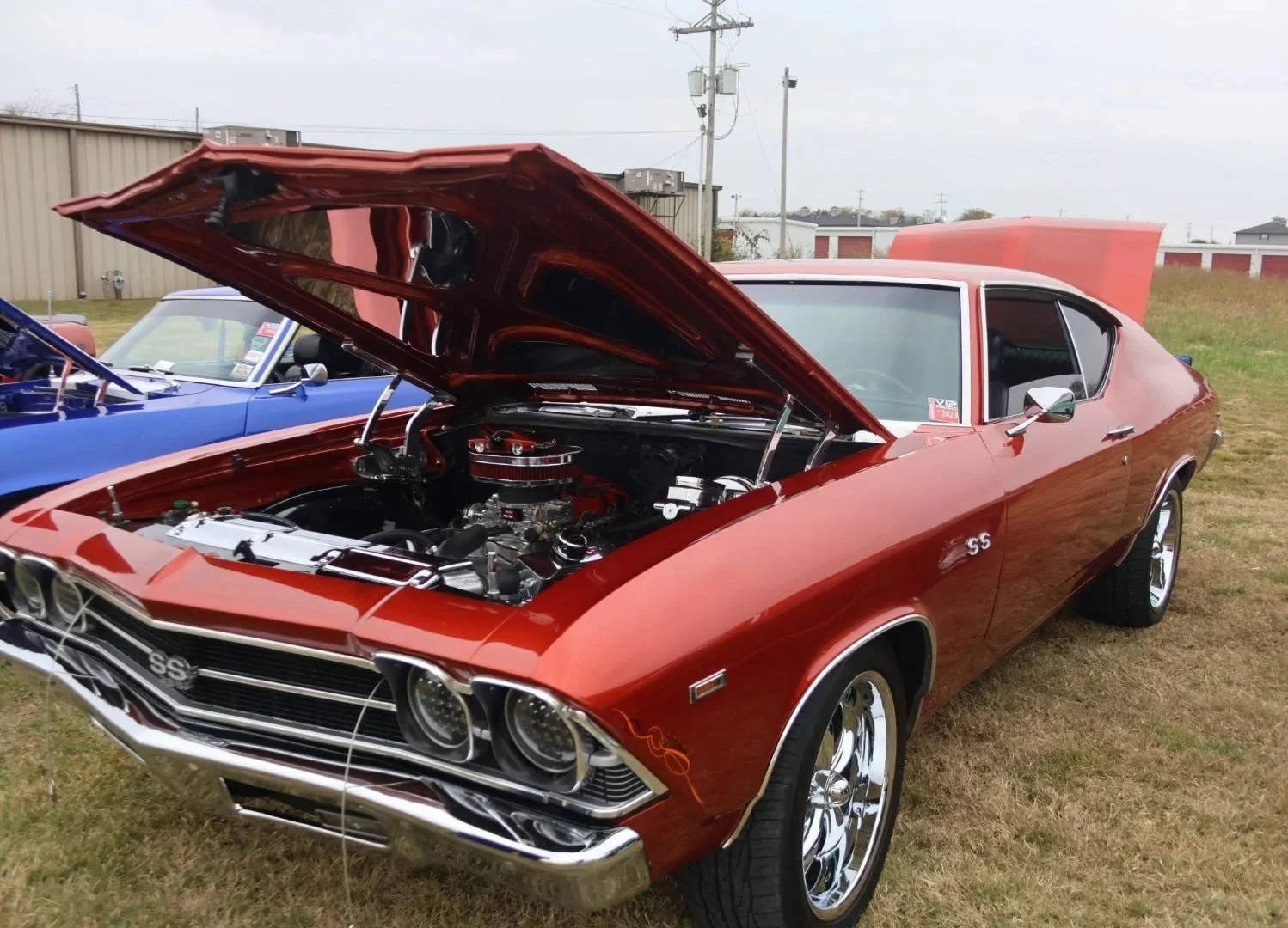 Red classic Chevrolet SS muscle car with its hood open, showing the engine at an outdoor car show, with a blue car nearby and buildings in the background.