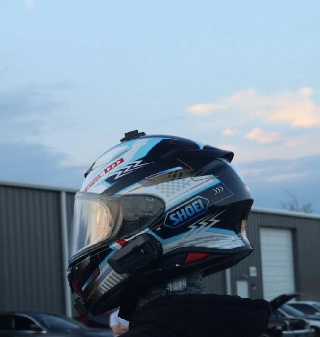Close-up of a motorcycle helmet with Shoei branding, black, blue, and white design, worn by a person in black clothing outside near a metal building.