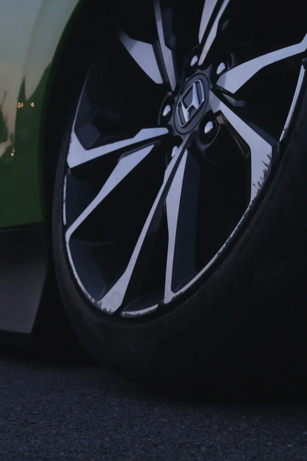 Close-up of a modern black and silver alloy wheel on a car, with a Honda logo at the center, parked on asphalt during evening or early morning.