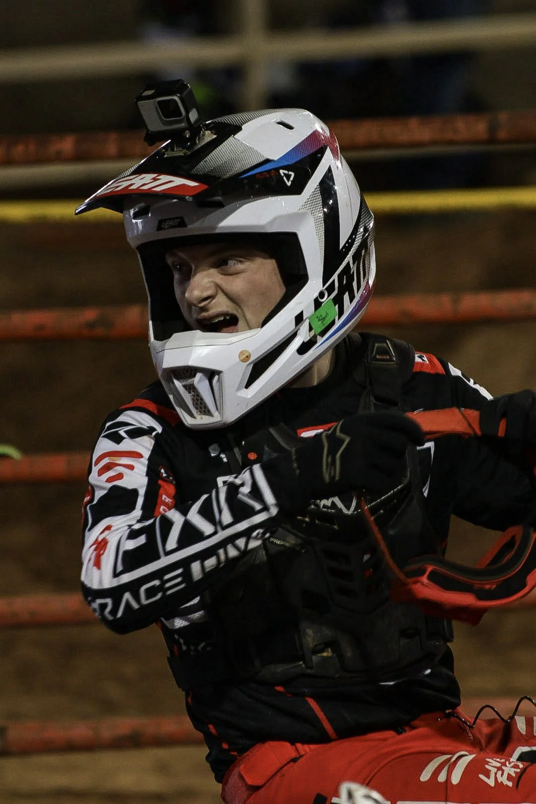 A person in motocross gear, including a white helmet, black and red racing suit, and red pants, riding a dirt bike on a rough track at night.