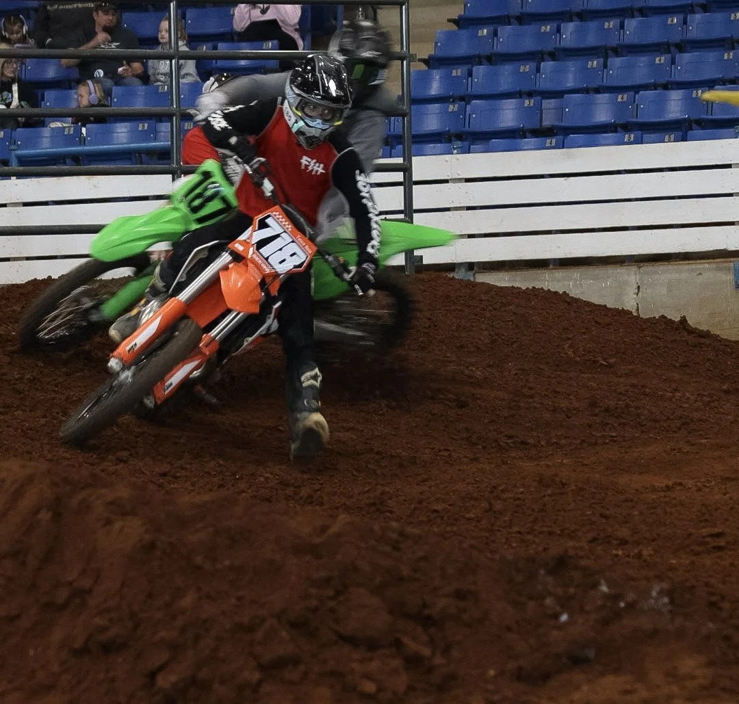 Motocross rider in black and red gear leaning into a turn on an orange motorcycle with number 718, on a dirt track with a green bike in the background and spectators in the bleachers.