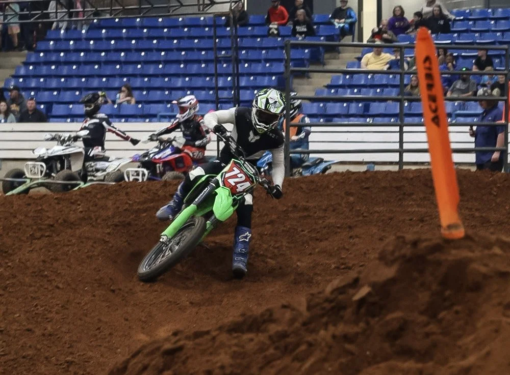 A motocross rider in black and white gear riding a green dirt bike on an indoor dirt track, with spectators seated in the background.