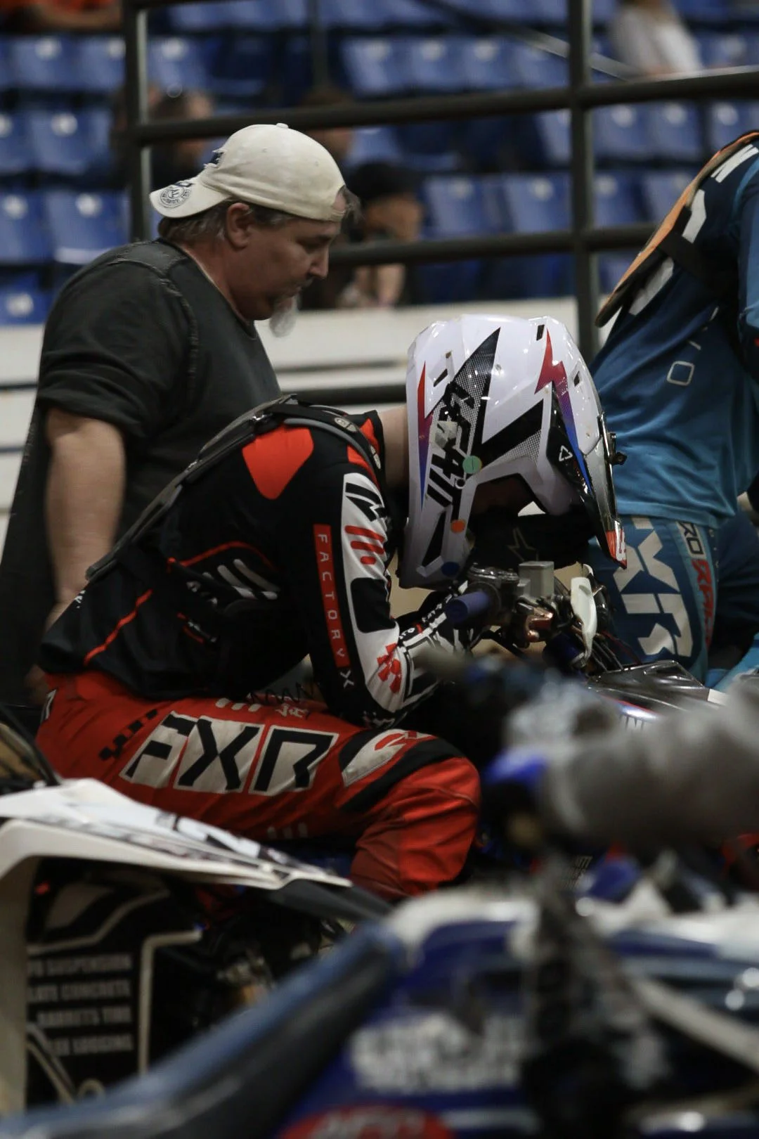 A motorcycle racer in red and black gear sitting on a motorcycle, with two men standing nearby, one wearing a beige cap and the other in a blue jacket, in a garage or paddock area.