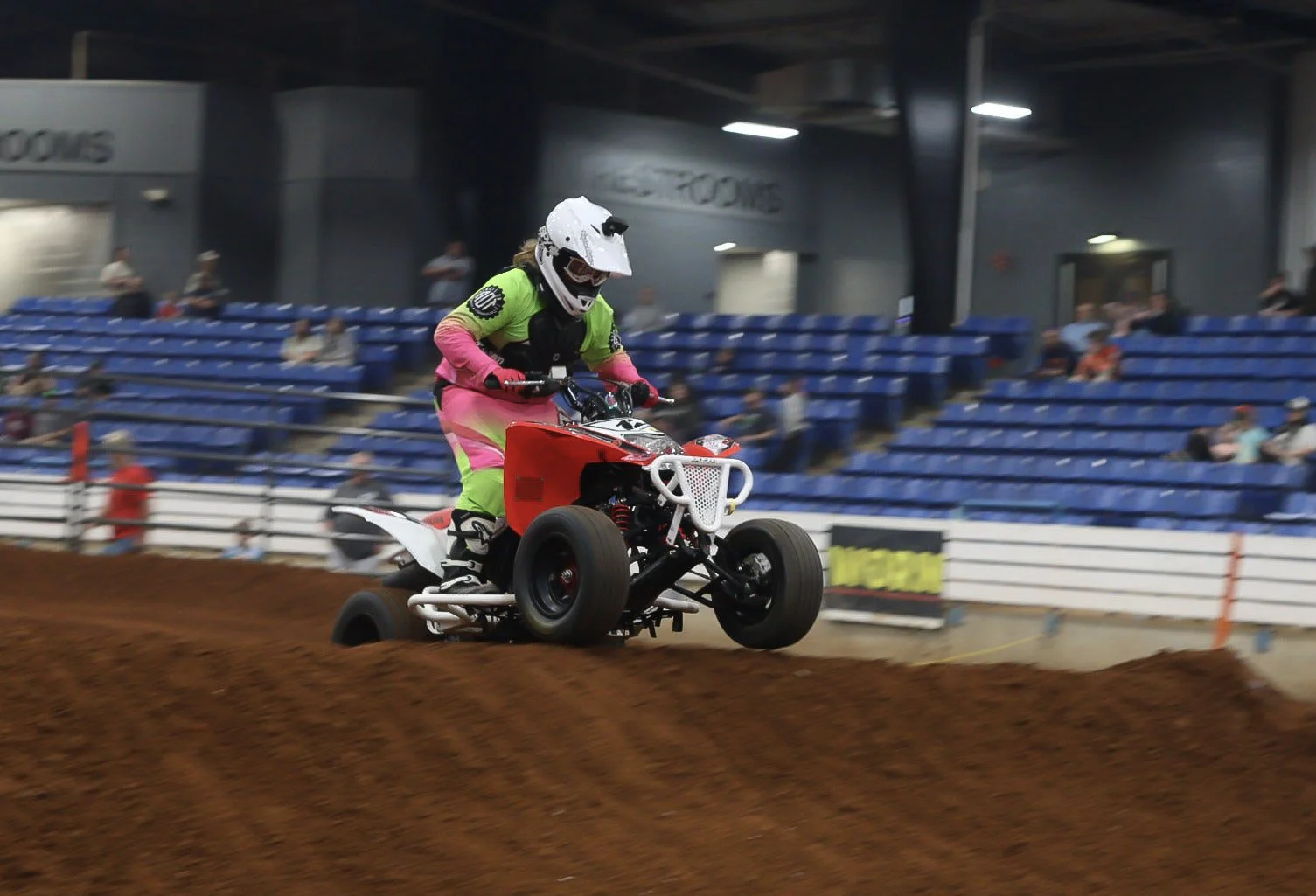A person riding a quad bike on an indoor dirt track, wearing a full-face helmet and colorful protective gear, with spectators seated in the background.