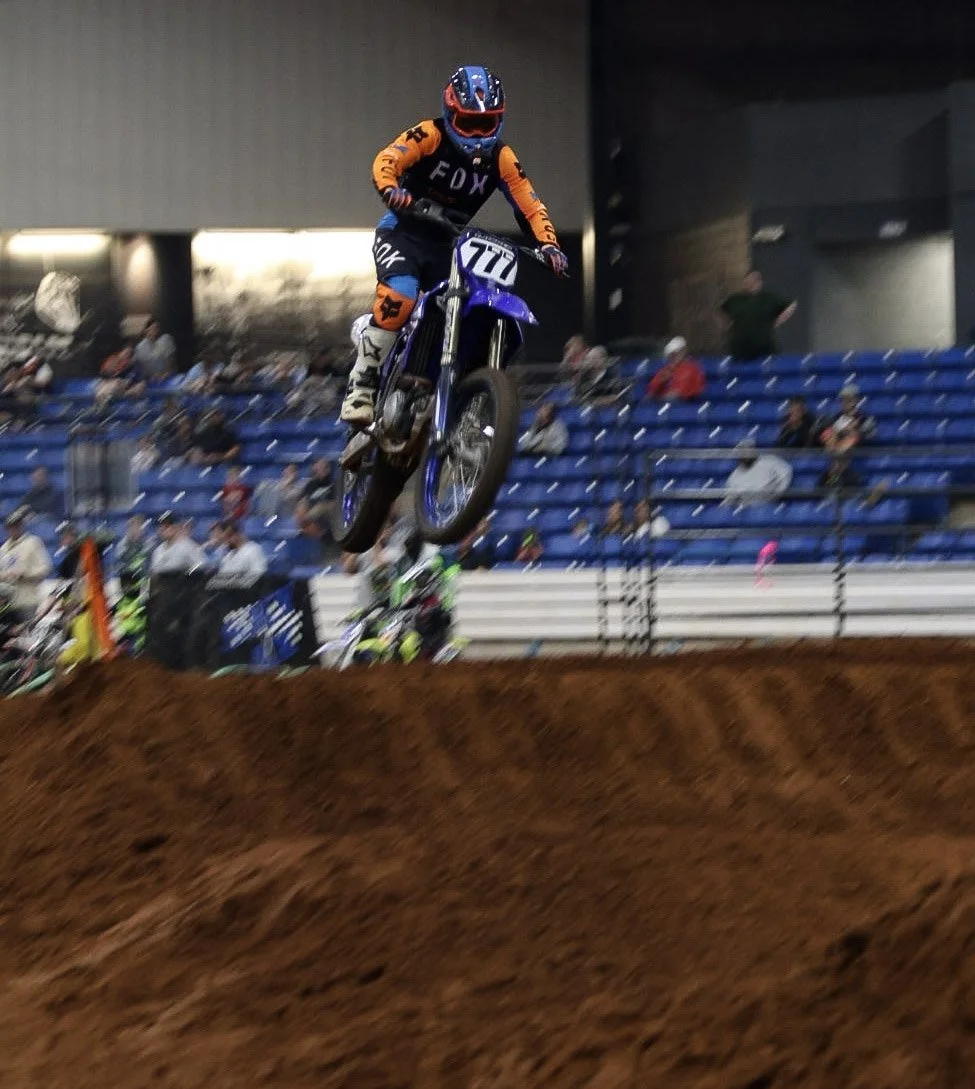 Motocross rider mid-jump in an indoor stadium, wearing orange and black gear with a blue helmet, on a blue and white dirt bike, with spectators in seats in the background.