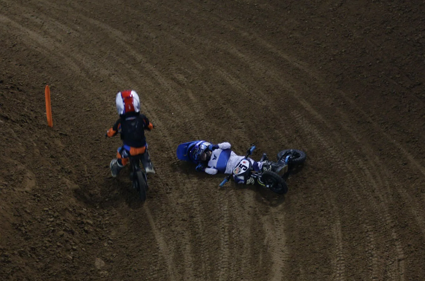 A dirt bike rider in white and blue gear lying on the ground after an accident on a dirt track, with another rider in orange and black riding past.
