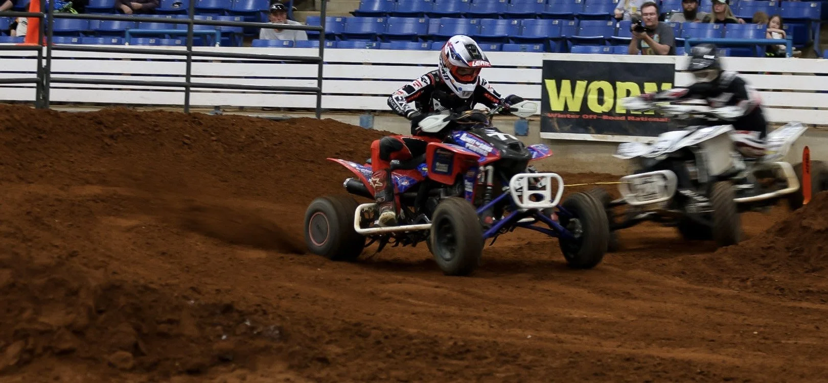 Two ATV racers competing on a dirt track, one rider in a red, black, and white suit with a helmet, the other in a black and white suit with a helmet, with an audience watching from the stands in the background.