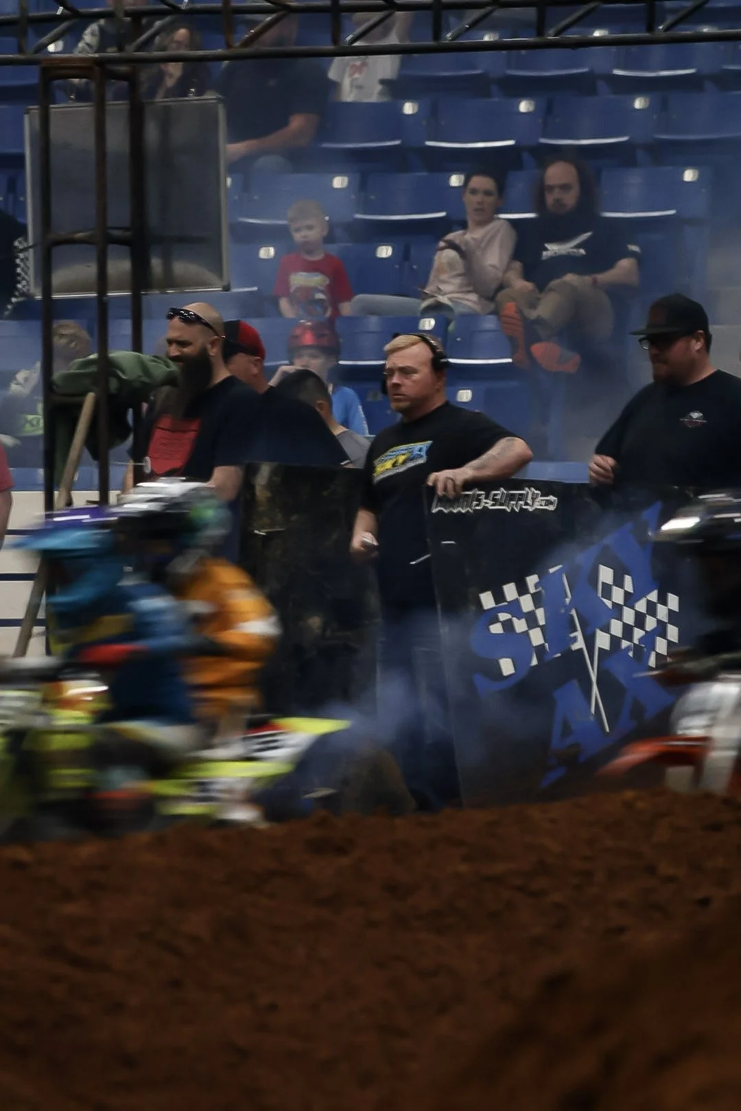 Motorcycle race track with riders in motion and spectators watching from the stands, with some seated and others standing.