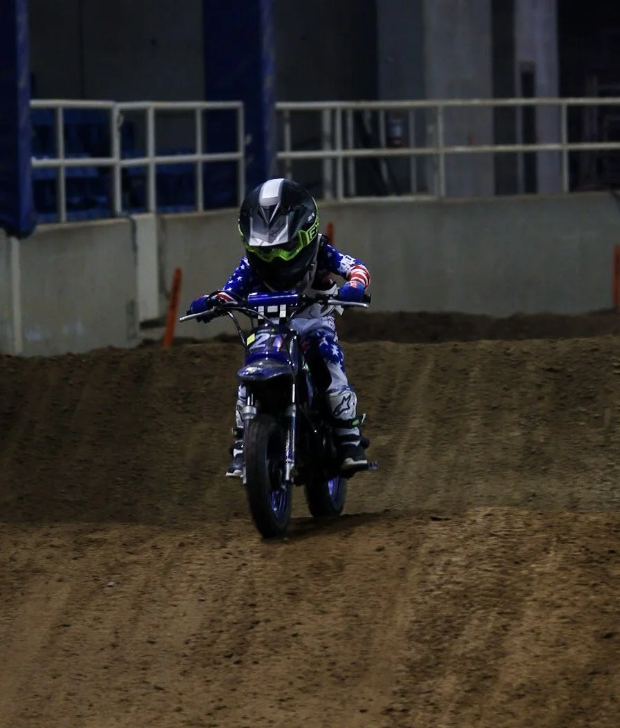 A young person riding a dirt bike on an indoor BMX track, wearing a black helmet with green accents and a patriotic motocross suit.