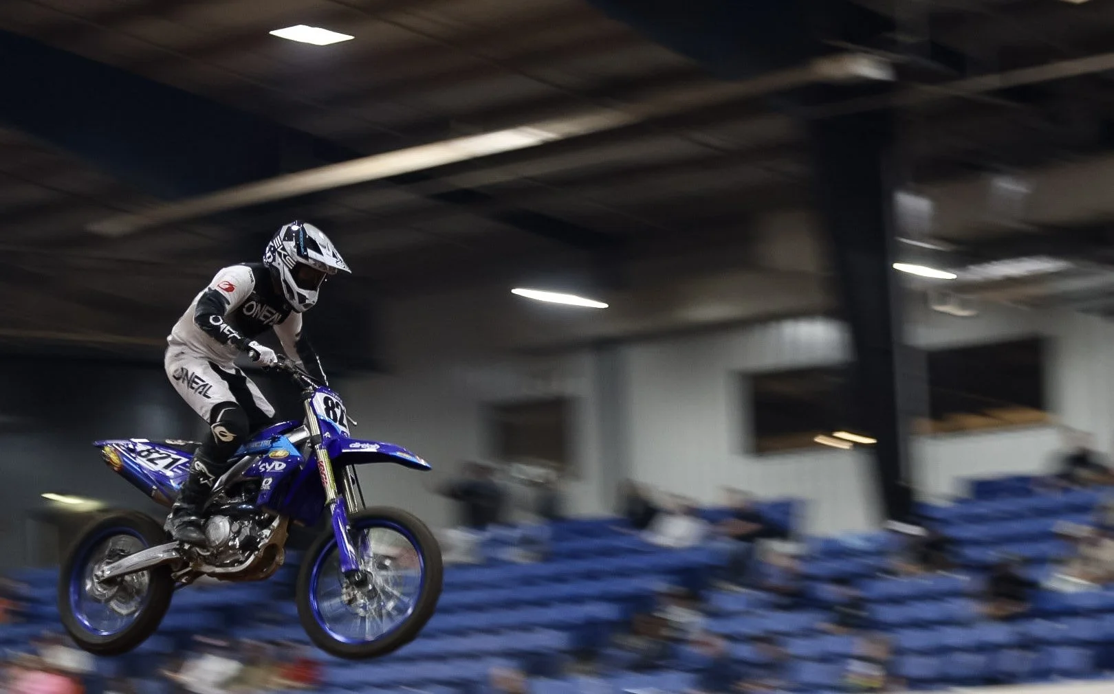 A motocross rider in white and black gear and helmet is mid-air on a blue dirt bike, racing indoors with blurred blue seating in the background.
