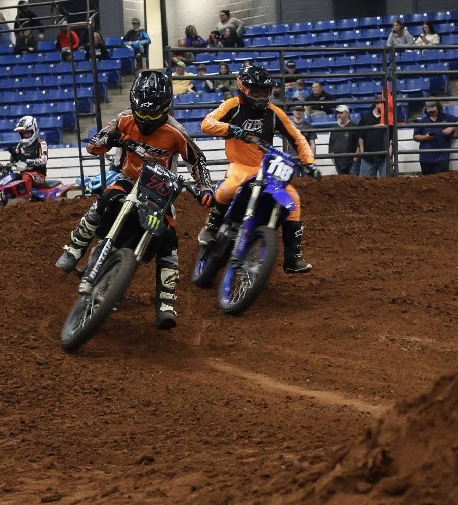 Two young motocross riders in orange and black gear riding dirt bikes on a track inside an indoor arena with blue spectator seats and people watching.