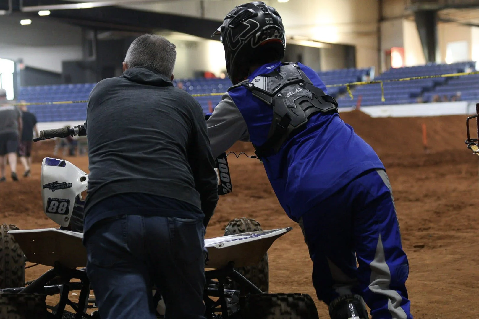Two individuals are working on a dirt bike inside an indoor motocross track. One person is facing away from the camera, wearing dark clothing, while the other is wearing blue motocross gear and a helmet, labeled 'Atlas'. The focus is on the bike and 