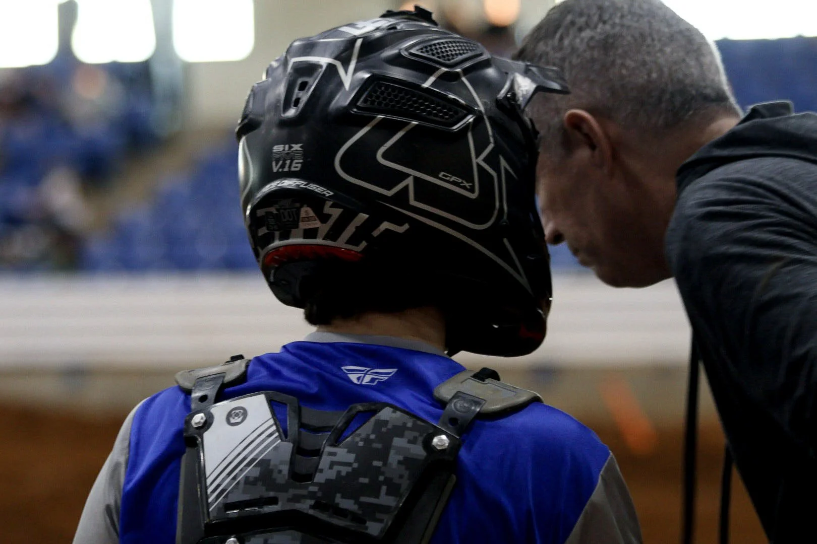 A motocross rider in blue gear and a black helmet with graphics, talking to a man in a black jacket at an indoor motocross event.