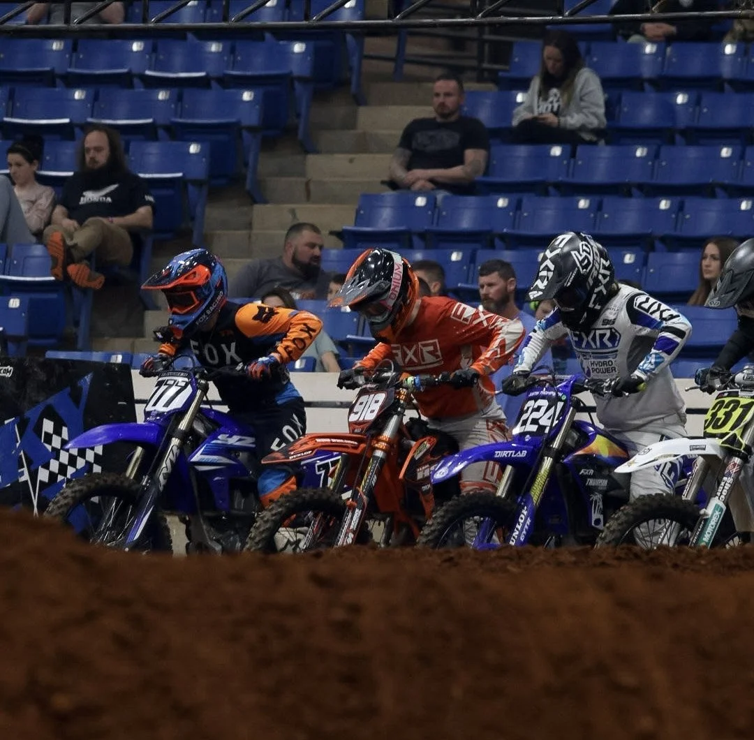 Motocross riders on their bikes preparing for a race in an indoor stadium with spectators in the background.