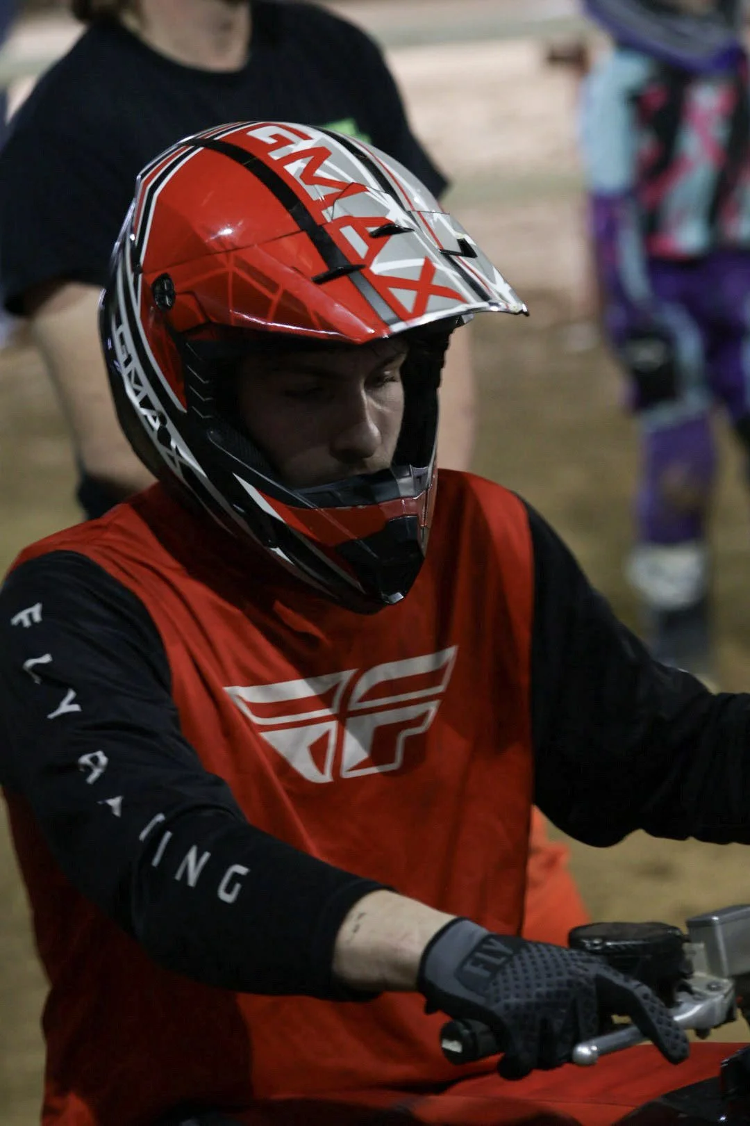 A person wearing a red and black motocross helmet and red motocross jersey with a logo, sitting on a motorcycle, indoors. The background shows other people, some wearing colorful clothing.