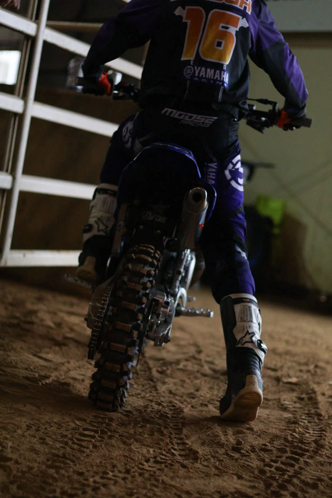 A motocross rider in racing gear riding a dirt bike indoors, with dirt floor and storage shelves nearby.