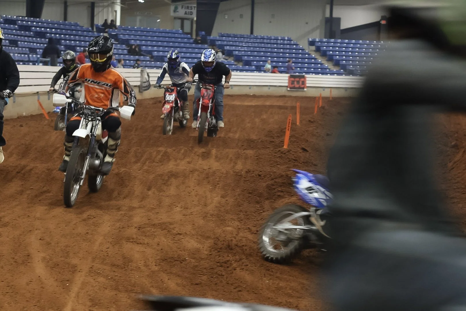 Indoor dirt bike track with riders wearing helmets and riding gear, some on motorcycles and one on a dirt bike, with blue stadium seating in the background.