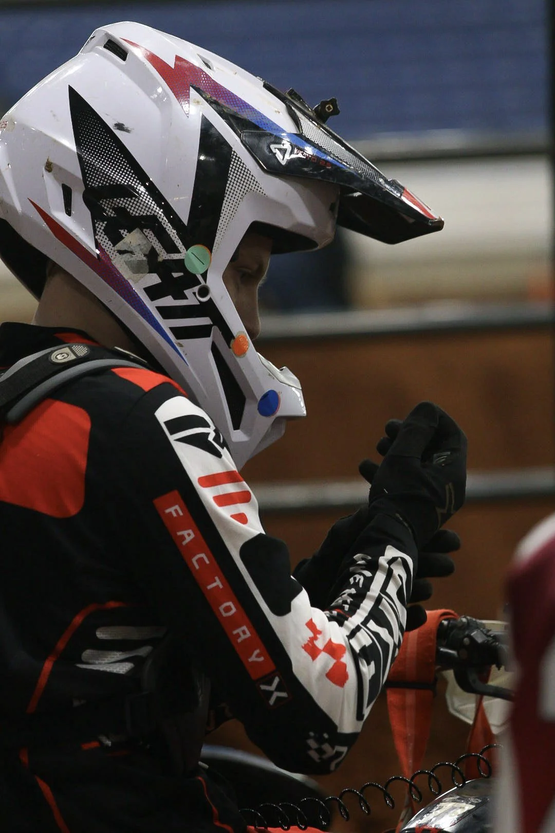 A person wearing a white and black motocross helmet and red, black, and white racing gear, standing next to a motorcycle with gloves on, in a racing setting.