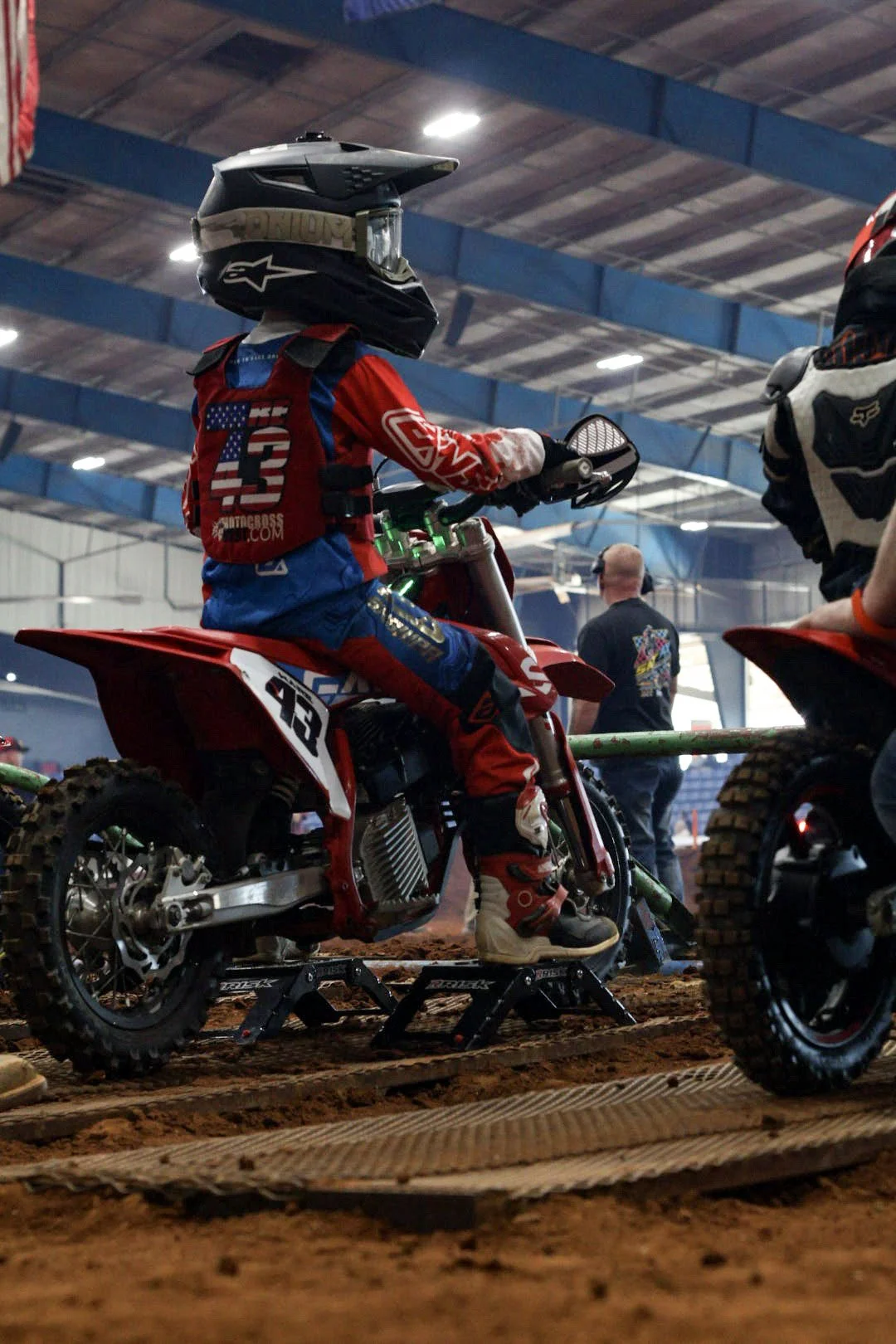 A young person wearing a motocross helmet, red and blue motocross gear with the American flag and number 13, sitting on a red dirt bike with the number 123, inside an indoor motocross arena with dirt floor and other riders in the background.