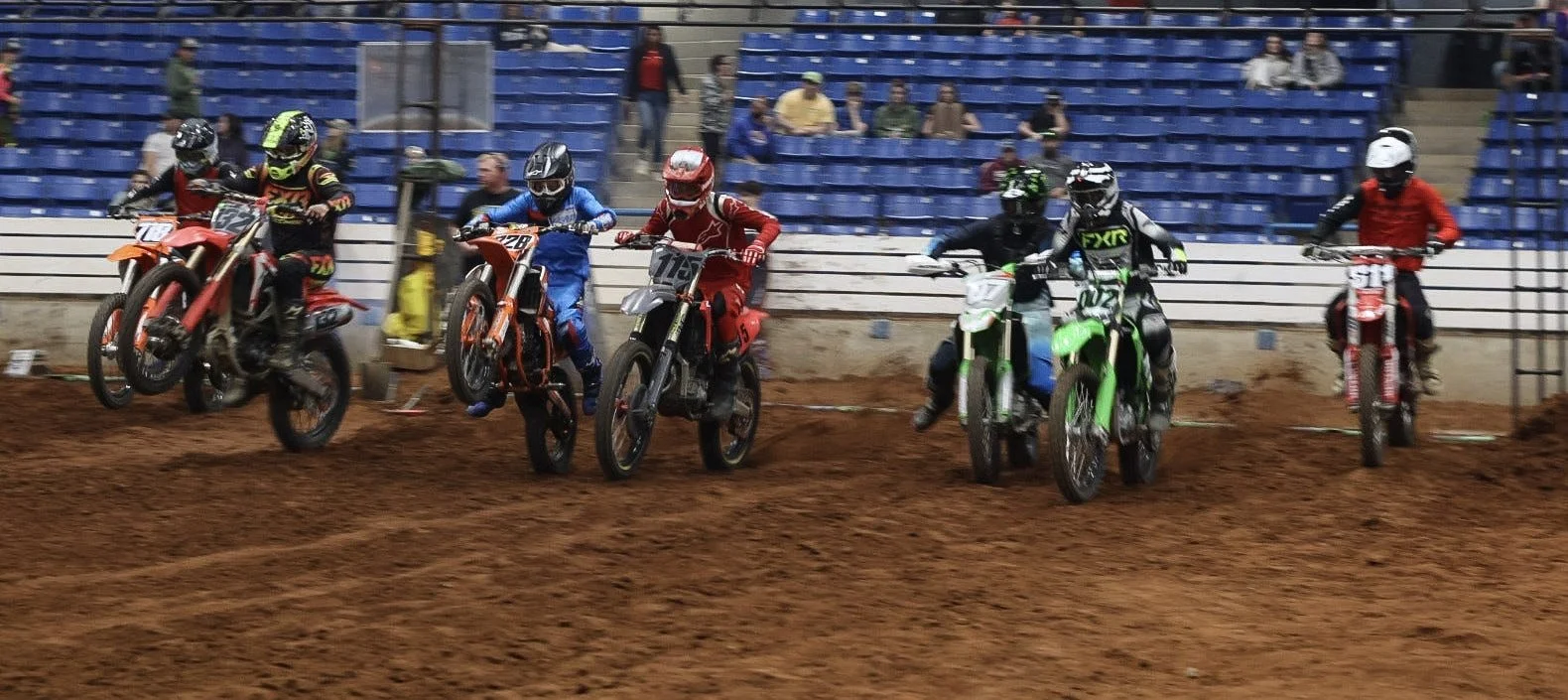Six motocross racers are starting a race on a dirt track, with some riders airborne as they accelerate. Spectators sit in the blue stands behind a fence.