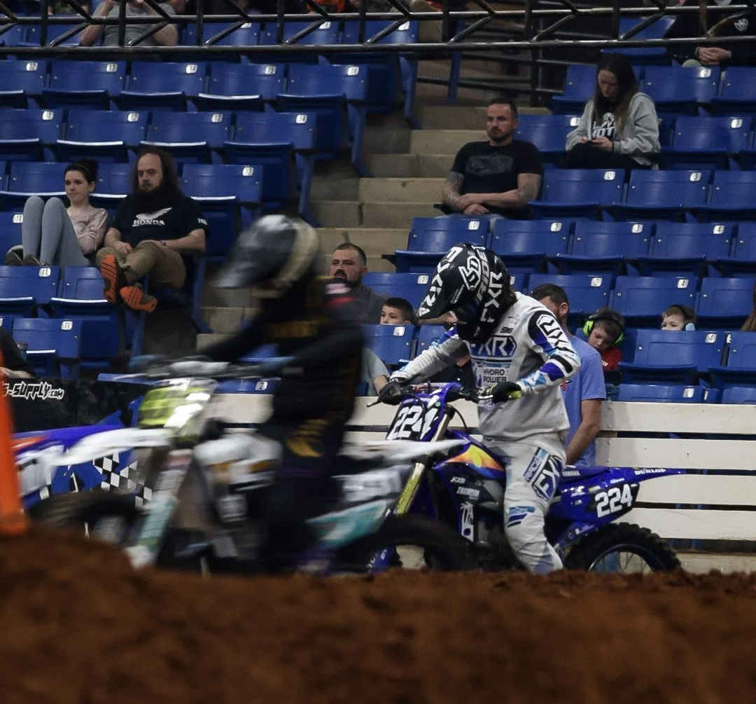 Motocross riders racing on an indoor dirt track, with spectators seated in blue bleachers watching.