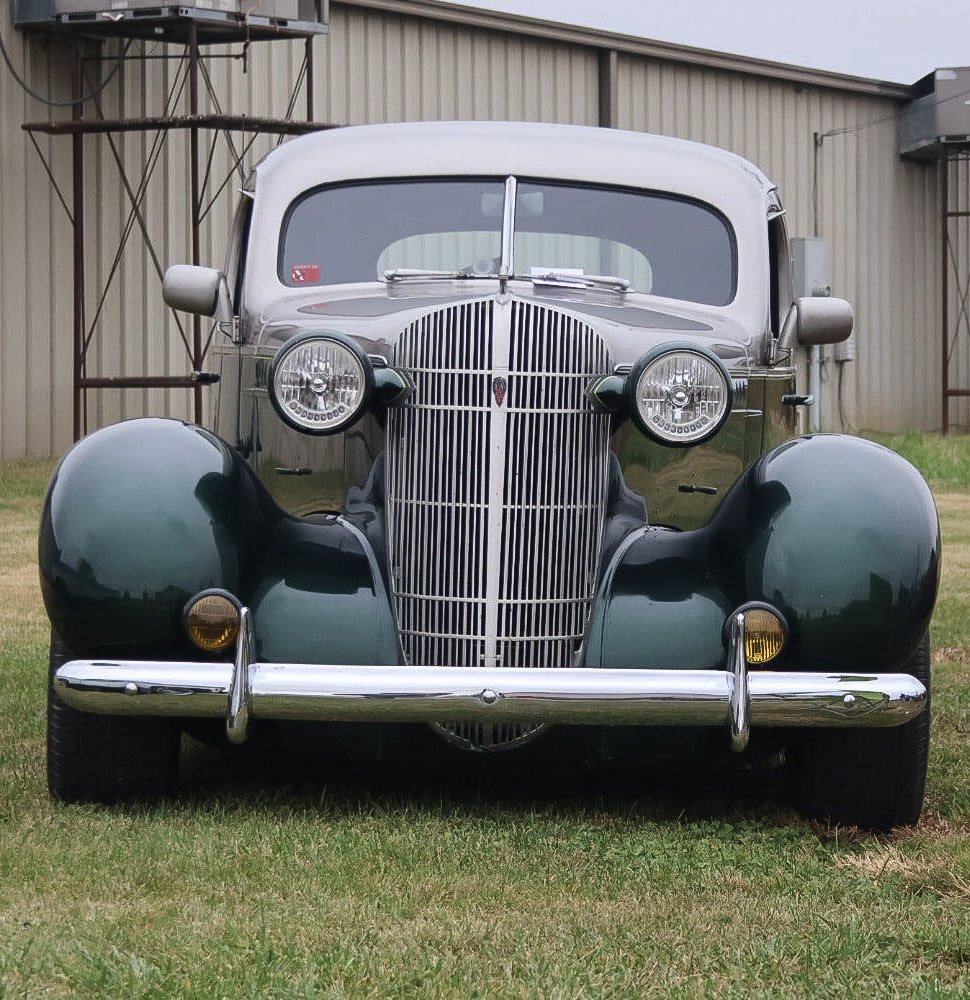 Front view of a vintage car parked on grass in front of a metal building, featuring prominent rounded fenders, a vertical grille, and round headlights.