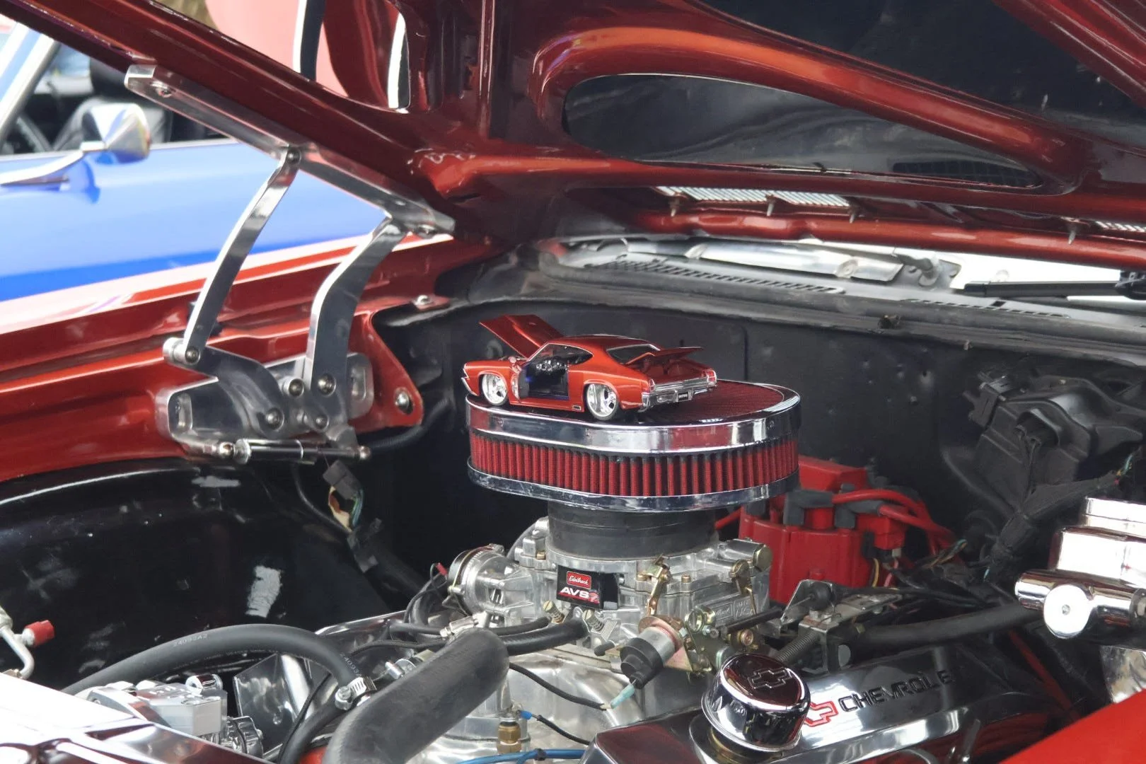 View of a classic car engine in a vintage Chevrolet car with a miniature model car on top of the air filter. The engine bay has chrome and red accents.