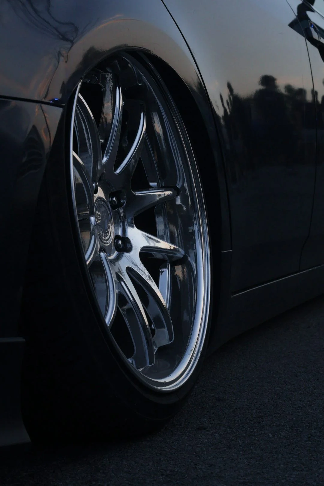 Close-up of a black car wheel with chrome rim and low profile tire, reflecting the surroundings, during dusk or evening.