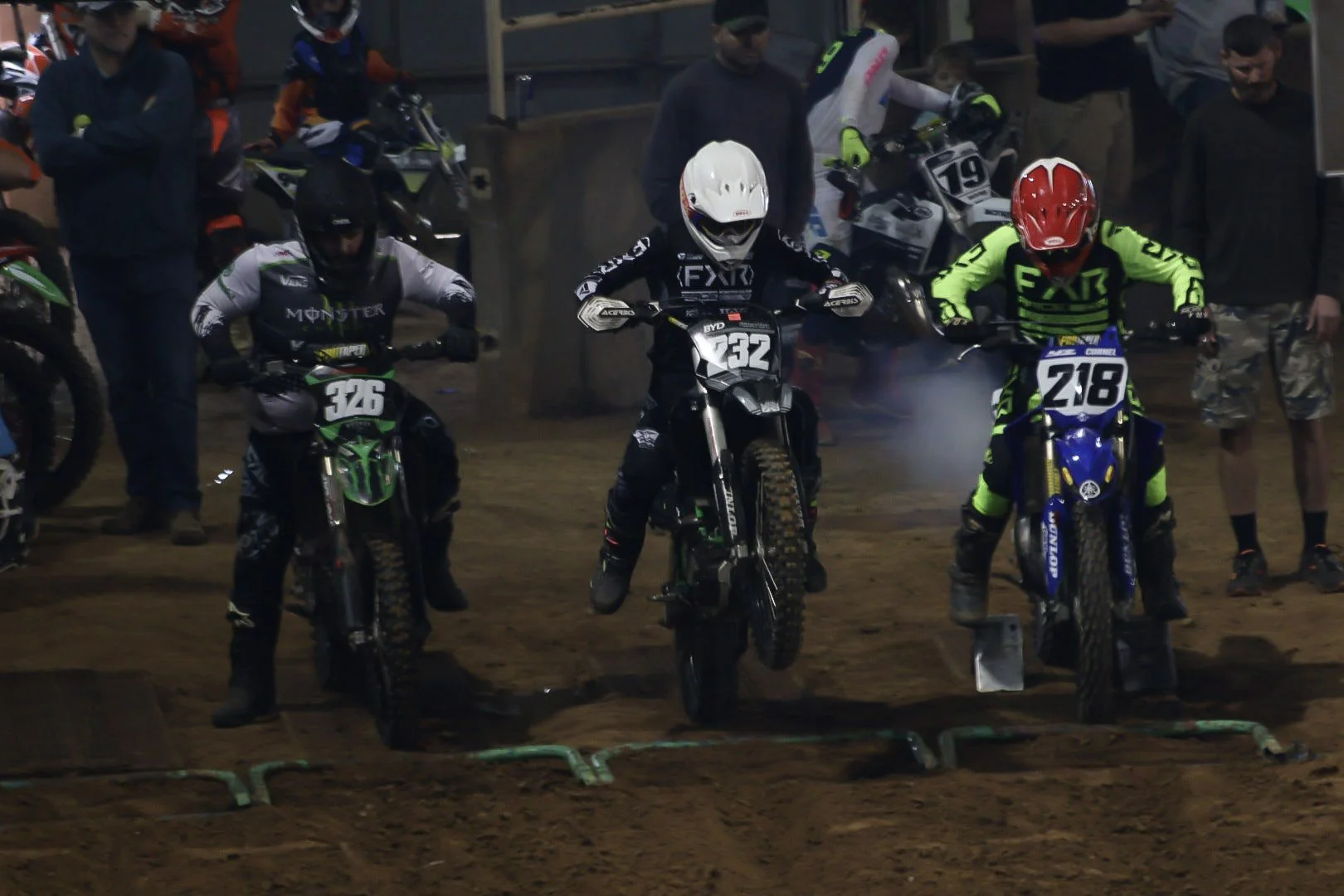 Motocross racers in gear on dirt bikes at the starting line, surrounded by spectators and crew.