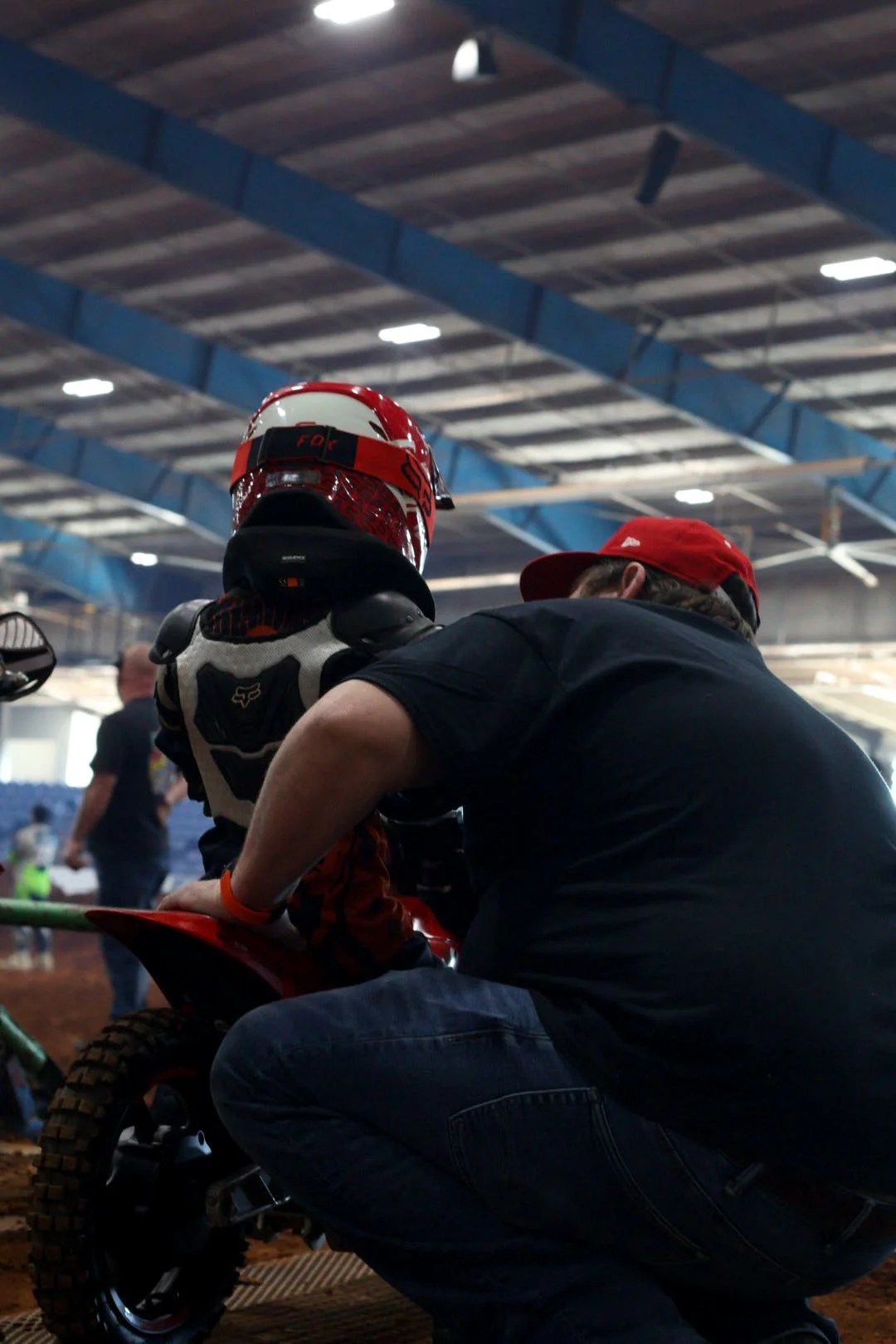 A person wearing a racing helmet and suit sits on a small dirt bike while another individual, wearing a black shirt and red cap, kneels beside them, possibly providing assistance or instructions, inside an indoor racing arena.