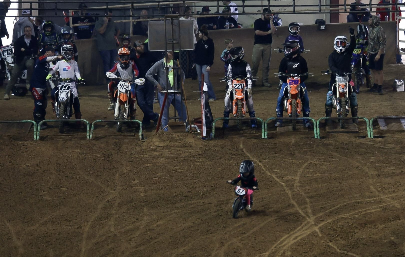 A young child riding a small pink bicycle with the number 67 on the front, riding on dirt at a motocross track. In the background, there are several adult motocross riders in gear and helmets, with their bikes lined up along a barrier, and spectators