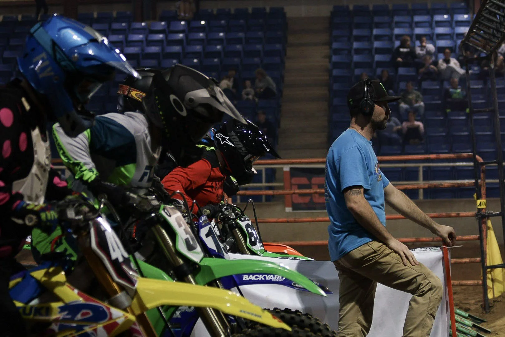 Motocross racers lined up on their bikes on the starting grid, with a crew member in a blue shirt and headset nearby, preparing for a race in an indoor arena with blue seating.